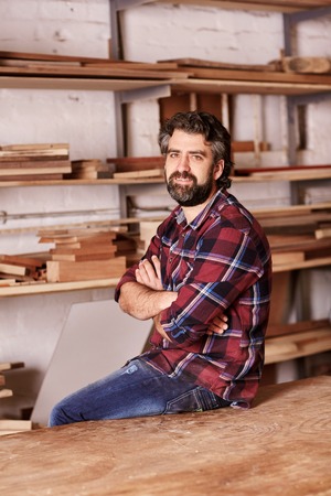Portrait of a small business owner sitting on the edge of his workbench, looking at the camera with his arms crossed confidently, with workshop shelves in the background with planks of woodの写真素材
