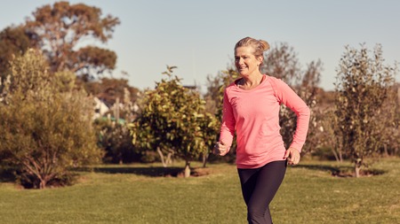 Smiling senior woman outdoors jogging, with grass and trees around her on a sunny morning, looking fit and healthyの写真素材