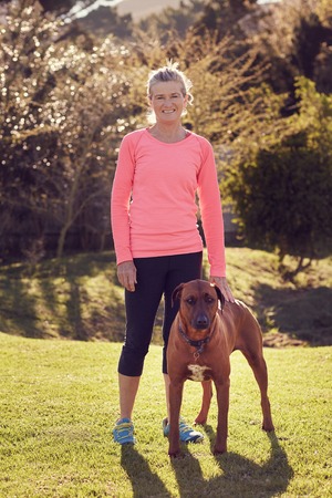 Full length image of a smiling senior woman wearing athletic clothing, standing outdoors on green grass with trees in the background and gentle sunlight, with her hand resting on the back of her dogの写真素材