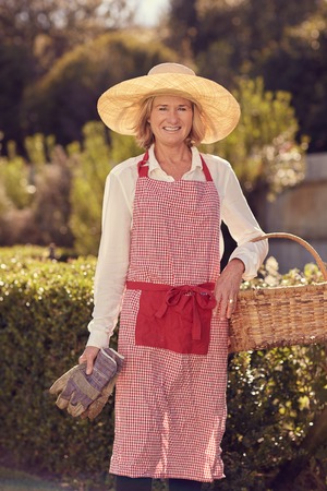 Portrait of a senior woman smiling at the camera while standing outdoors in front of trees and a hedge wearing a straw hat and apron, and holding gloves and a basket for gardeningの写真素材