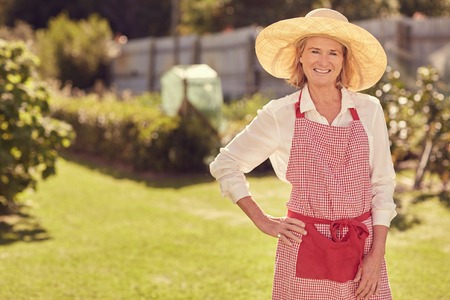 Portrait of a senior woman in hat and apron, smiling at the camera with her hand on her hip, on a summer day in her lush backyard gardenの写真素材