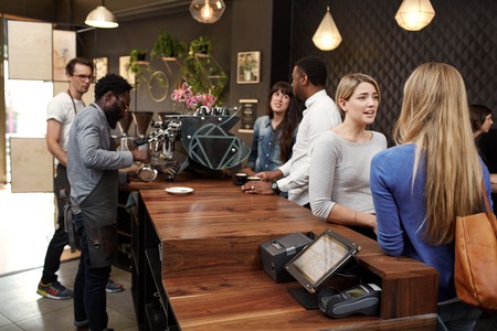 Mixed racial group of people in a modern cafe, baristas working behind the wooden counter and customers chatting while waiting for their coffees to be madeの写真素材