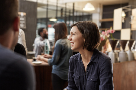 Candid shot of a woman with bobbed brunette hair looking away at someone out of frame and smiling broadly and with confidence, while customers are being served in the backgroundの写真素材