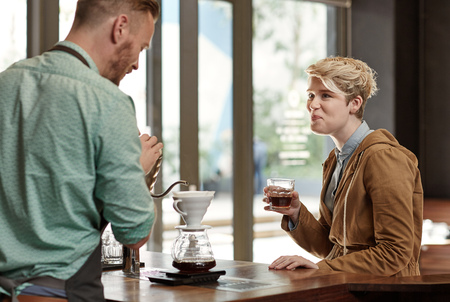 Friendly barista busy pouring a fresh cup of filter coffee while chatting to a regular young woman customer, who is enjoying a glass of filter coffee on a work breakの写真素材