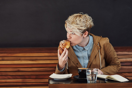 Young woman with trendy blonde hair looking away while taking a bite from her pastry, at a table in a modern cafe with a dark grey wallの写真素材