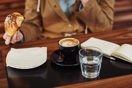 Cropped image of a person sitting at a modern cafe holding the croissant they are eating, with an open notebook, cappucino and a glass of water on the tableの写真素材
