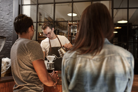 Barista carefully pouring coffee through a special filter at a modern coffee shop with two customers...