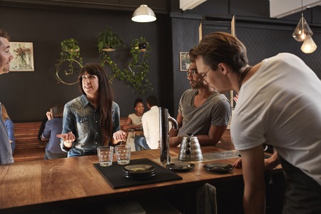 Woman chatting with colleagues at the wooden counter of a trendy, hipster style coffee shop while on...