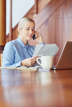 A young businesswoman at her desk, talking on phone while checking documents, inside wooden officeの写真素材