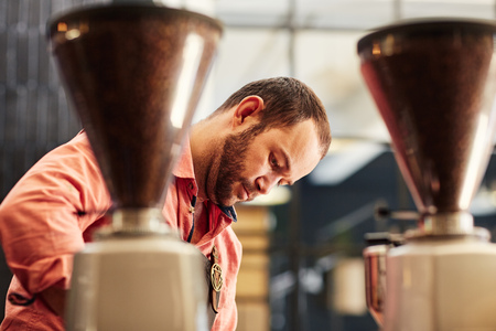 Caucasian man working in a coffee roastery, concentrating on his work behind two modern coffee gindersの写真素材