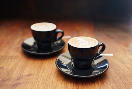 Modern black ceramic cups and saucers containing freshly made cappucinos, sitting on a wooden counterの写真素材