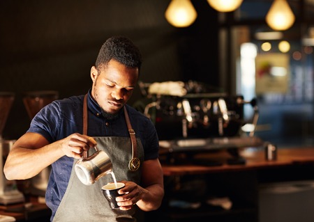 Handsome African man pouring frothed milk carefully into a cappucino, with a gentle morning light and a modern coffee shop behind him where he has a job as a baristaの写真素材