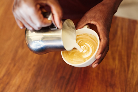 Cropped closeup of the hands of an african barista carefully pouring foamed milk from a stainless stell jug, into a coffee cup to form a fancy pattern on a cappucinoの写真素材
