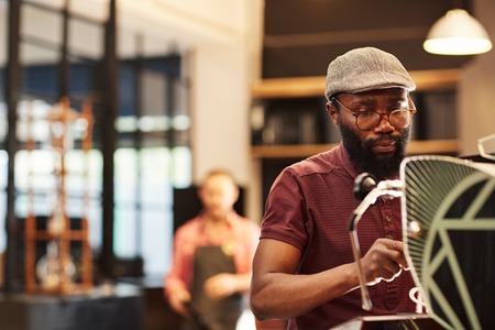 Handsome Afro-American man in hipster cap and spectacles, making coffee at an espresso machine in a...