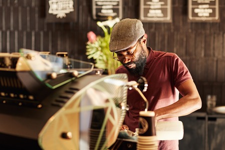 Handsome African man with a hipster style, working behind an espresso machine in a modern coffee shop where he has a job as a baristaの写真素材
