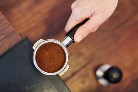 Overhead shot of a person's hand holding the handle of shiny new portafilter for an espresso machine containing a perfectly level amount of fresh ground coffee, with a tamper visible on counter belowの写真素材