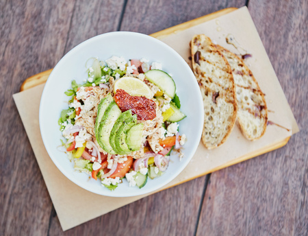Overhead shot of a fresh salad consisting of chicken, avocado, sundried tomatoes and other tasty ingredients on a wooden table with freshly sliced bread alongsideの写真素材