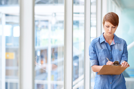 Female intern wearing a casual denim shirt writing notes on a piece of paper on a clipboard, while standing in a corridor with large windows and lots of lightの写真素材