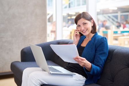 Attractive young businesswoman sitting on an office couch smiling at the camera, while holding some paperwork and her phone to her ear, with her laptop computer on her lapの写真素材