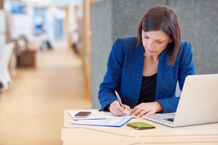 Stylish young busineswoman writing while working on paperwork at her desk in a bright shared office with a cucbicle divider behind herの写真素材