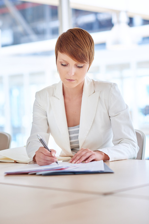 Pretty young businesswoman with a modern short hairstyle, sitting at a desk in a bright office, looking serious while writing on some paperworkの写真素材