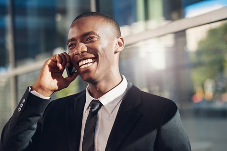 Handsome young businessman of African descent wearing a smart suit, holding his mobile phone to his ear and laughing with a friendly smile, while standing outdoors in a cityの写真素材