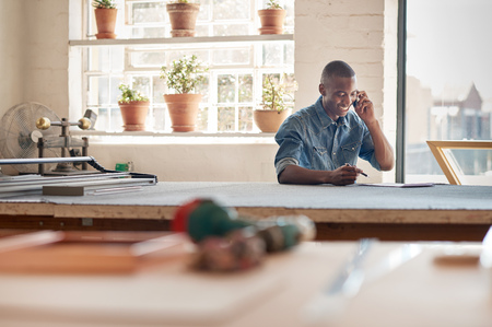 Handsome young African designer sitting in his workshop and smiling, while talking on his mobile phone discussing work related to his entrepreneurial businessの写真素材