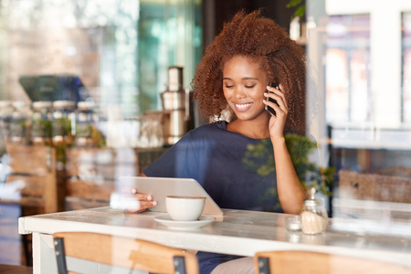 Attractive young woman using a digital tablet and talking on the phone while sitting in a cafeの写真素材