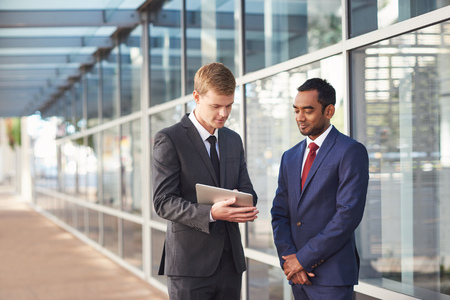 Two confident and successful businessmen talking together over a digital tablet while standing in front of glass windows of a modern office buildingの写真素材