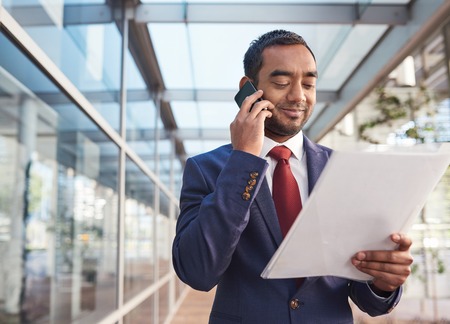 Confident and successful businessman talking on a cellphone and reading paperwork while standing in front of the glass windows of a modern office buildingの写真素材
