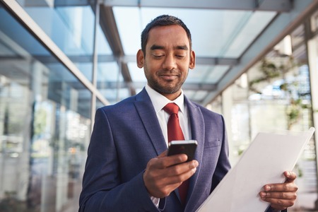 Confident and successful businessman texting on a cellphone while standing in front of the glass windows of a modern office buildingの写真素材