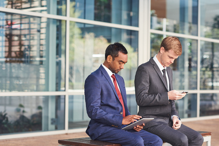 Two confident and successful businessmen sitting in front of the glass windows of a modern office building using a digital tablet and cellphoneの写真素材