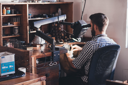 Jeweler sitting in his workshop looking through a large magnifier while doing precision work on a ringの写真素材