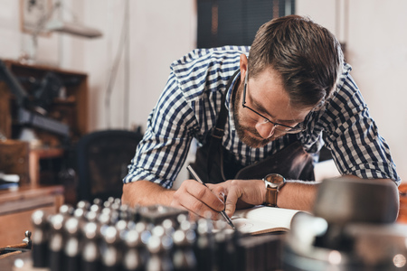 Jeweler leaning on a bench sketching out new jewelry designs in a notebook while working in his shopの写真素材