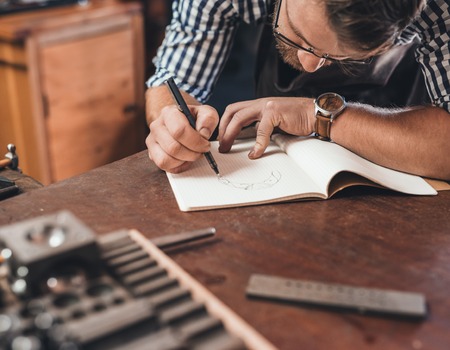 Jeweler leaning on a bench sketching out new jewelry designs in a notebook while working in his shopの写真素材