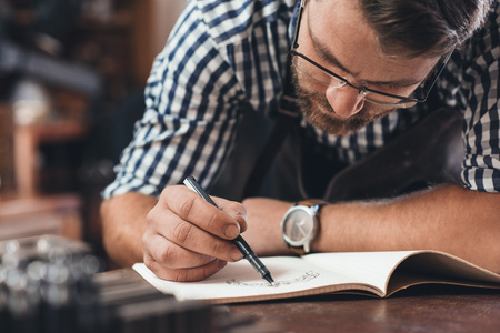 Closeup of a jeweler leaning on a bench sketching out new jewelry designs in a notebook while...