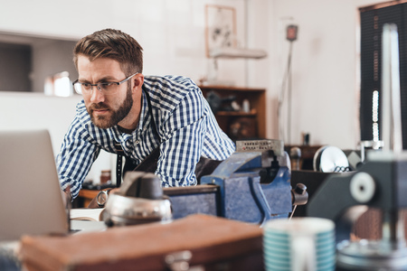 Focused young jeweler in an apron working on a laptop while leaning on a bench in his workshopの写真素材