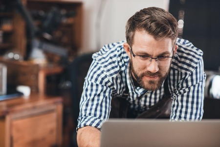Focused young jeweler in an apron working on a laptop while leaning on a bench in his workshopの写真素材
