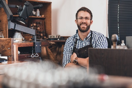 Portrait of a smiling young jeweler in an apron sitting at a bench full of tools in his workshopの写真素材