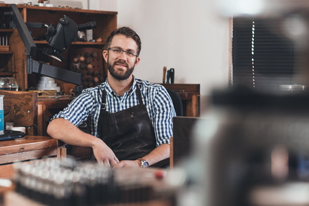 Portrait of a smiling young jeweler in an apron sitting at a bench full of tools in his workshop