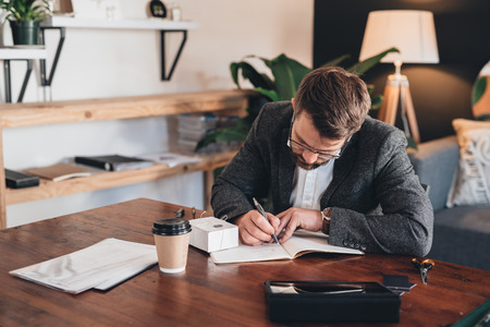 Young entrepreneur sitting at a table at home looking focused while writing in notebook next to a small boxの写真素材