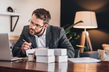 Young entrepreneur sitting at a table at home looking at a laptop and writing addresses on packages for delivery to customersの写真素材