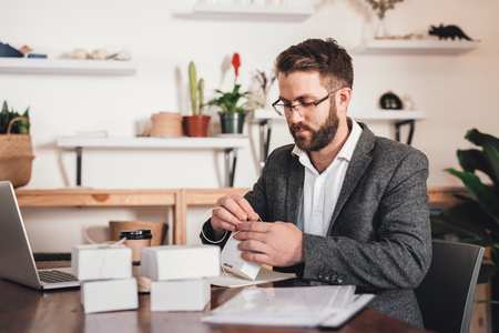 Focused young entrepreneur sitting at a table at home next to a laptop tying up packages for delivery to customersの写真素材