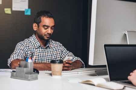 Designer sitting at his desk checking his cellphone and working on a computer while sitting in front of a colleague in an officeの写真素材