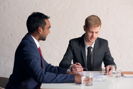 Two young businessmen in suits discussing documents together while sitting at a table in a modern boardroomの写真素材