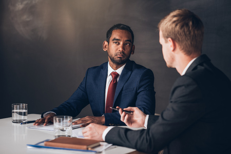 Two young businessmen in suits discussing documents together while sitting at a table in a modern boardroomの写真素材
