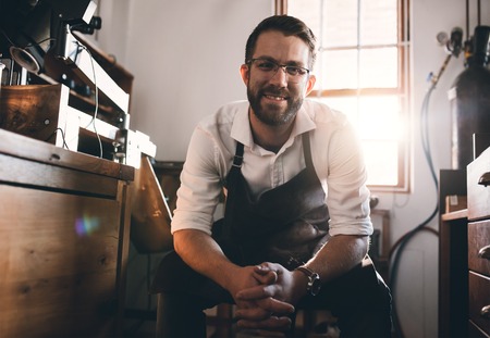 Portrait of a smiling young jeweler sitting in a workshop full of tools and jewelry making equipment