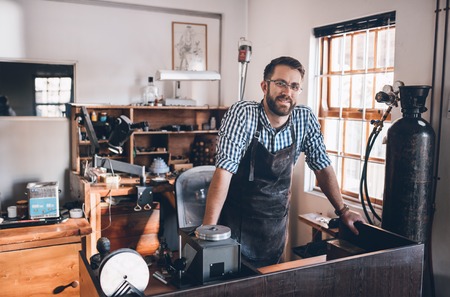 Portrait of a smiling young jeweler standing in a workshop full of tools and jewelry making...