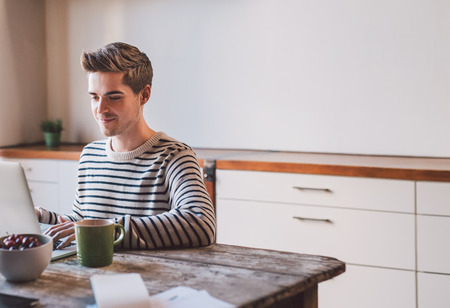 Smiling young man using a laptop while sitting at the kitchen table at homeの写真素材
