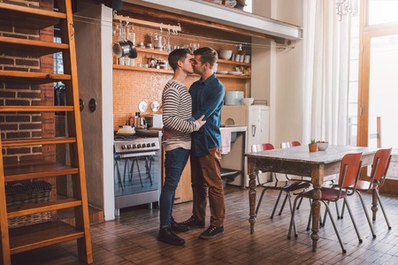 Affectionate young gay couple kissing while standing arm in arm together in their kitchenの写真素材